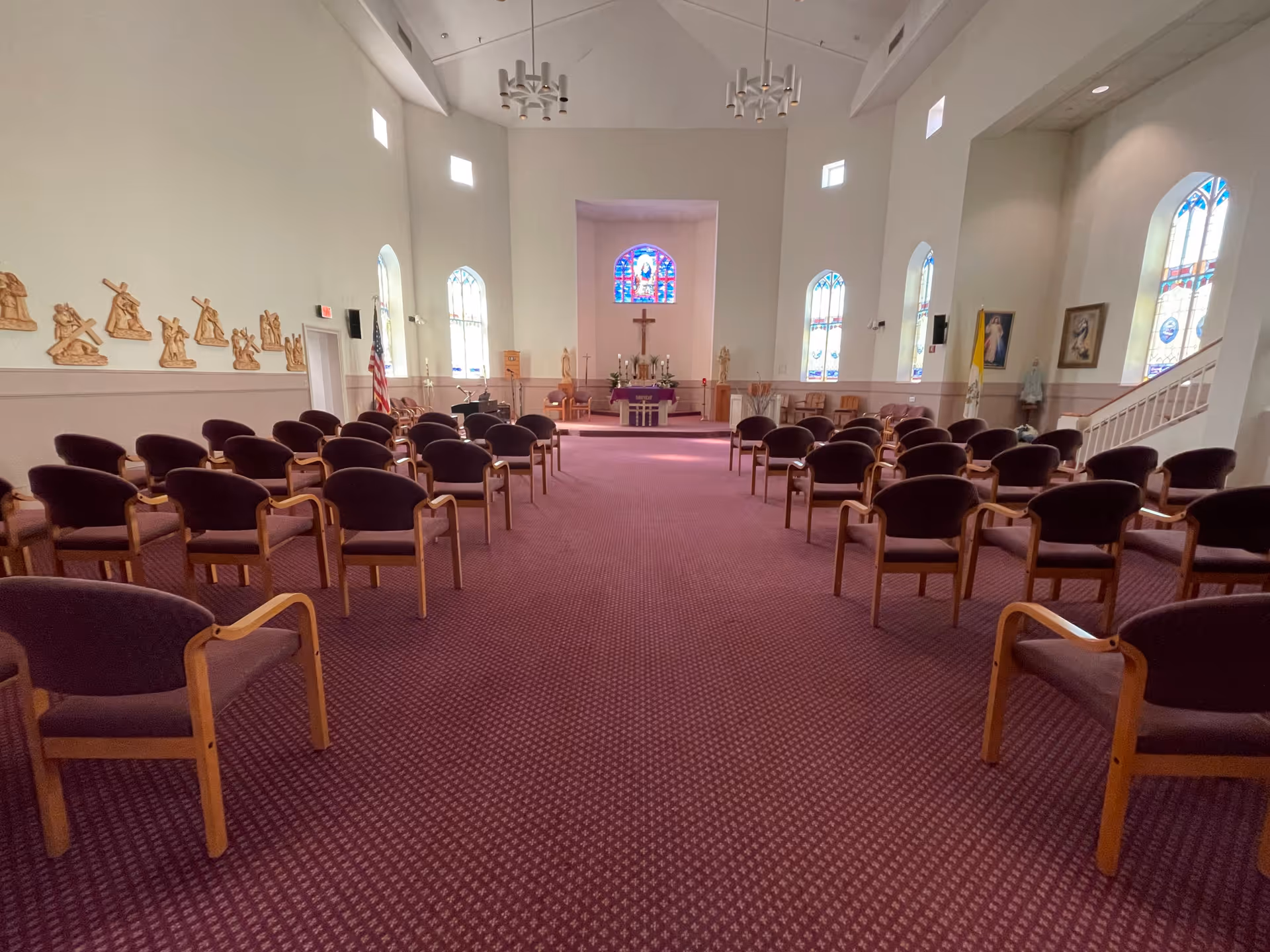 Interior view of a chapel with rows of purple cushioned chairs arranged facing an altar. The altar area features a cross, candles, and religious statues. The room has high ceilings, stained glass windows, and wall-mounted religious artwork.