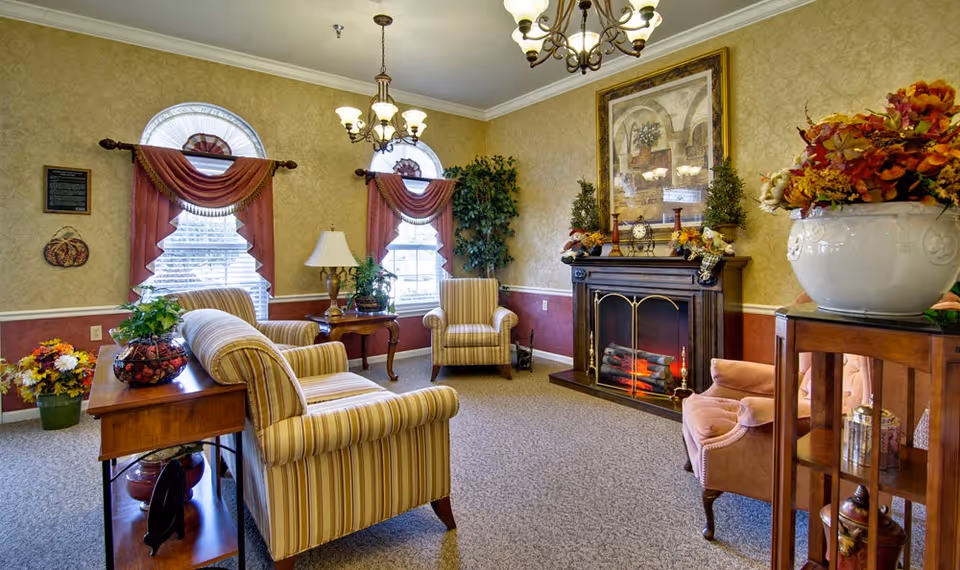 A cozy living room in a senior living facility with striped armchairs, a wooden side table with a lamp and plants, a fireplace decorated with autumn-themed ornaments, and a large framed painting above it. The room has two arched windows with red curtains and a chandelier hanging from the ceiling.