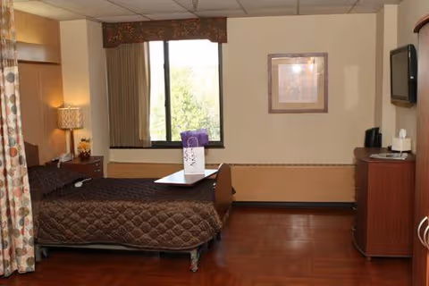 A senior living facility bedroom with a single bed covered in a brown quilted blanket. There is a bedside table with a lamp and a small decorative item. A window with curtains lets in natural light. On the right side, there is a wooden dresser with a TV mounted above it and a coffee maker on top. A framed picture hangs on the wall.