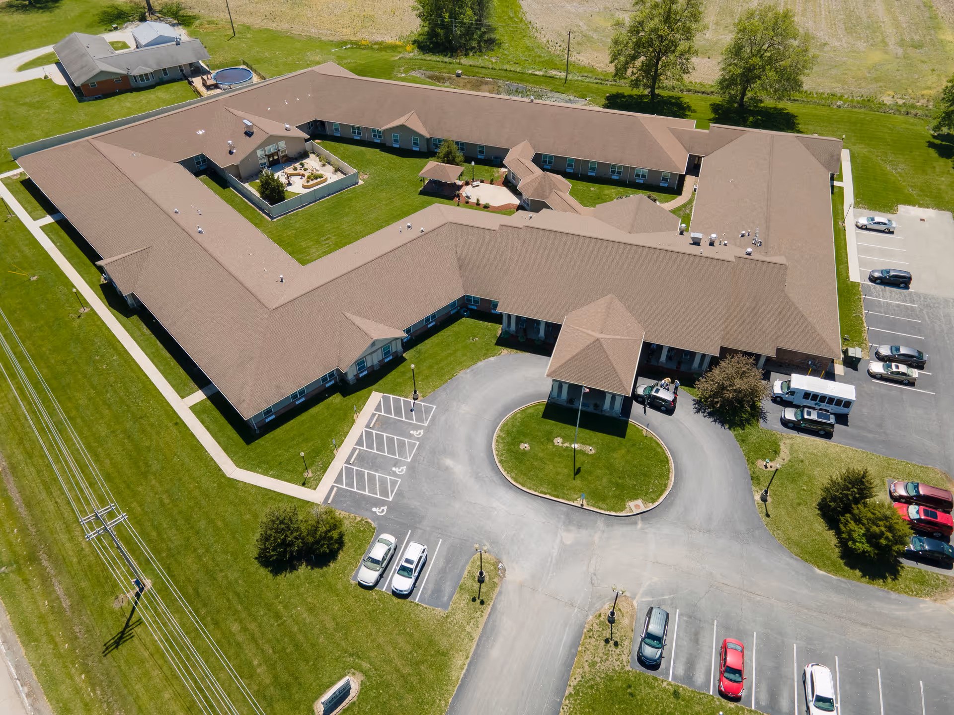 Aerial view of Cedar Trails Senior Living facility showing a large, single-story building with a brown roof arranged in a U-shape around a central courtyard. The building is surrounded by green lawns and parking lots with several cars parked. There is a circular driveway at the entrance with a covered drop-off area.