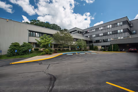 Exterior view of a multi-story nursing and rehabilitation center building with a parking area in front, surrounded by trees and bushes under a partly cloudy sky.