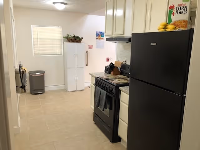 Small galley-style kitchen with a black refrigerator and stove, white cabinets, and a freestanding pantry cabinet at the far end.