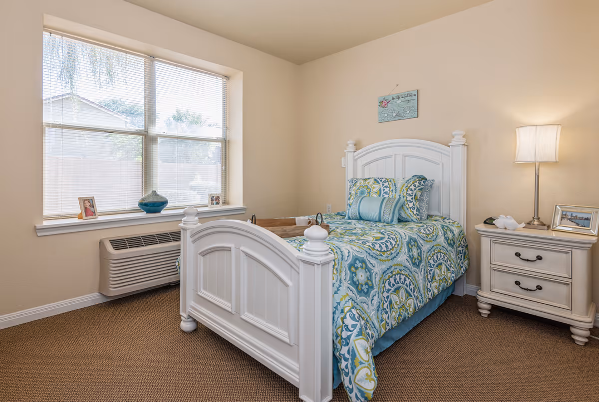 A cozy bedroom with a white wooden bed frame and a colorful patterned bedspread in shades of blue, green, and white. Next to the bed is a white nightstand with two drawers, a table lamp, and framed photos. A large window with blinds lets in natural light, and a heating/cooling unit is installed below the window. The walls are painted a light beige color.