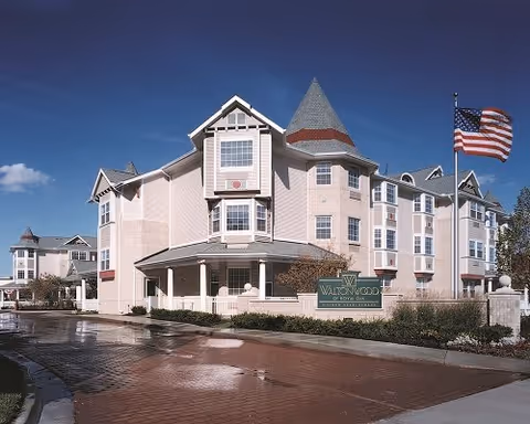 Exterior view of a multi-story senior living facility building with beige and white walls, multiple windows, and a turret-like architectural feature. An American flag is flying on a flagpole to the right, and there is a green sign in front of the building that reads 'Waltonwood Royal Oak'. The sky is clear and blue.