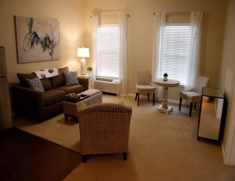 A cozy living room with a brown sofa adorned with patterned pillows and a throw blanket, a patterned armchair, a small ottoman, a side table with a lamp and plant, two windows with white blinds and sheer curtains, and a small round table with two chairs near the windows.