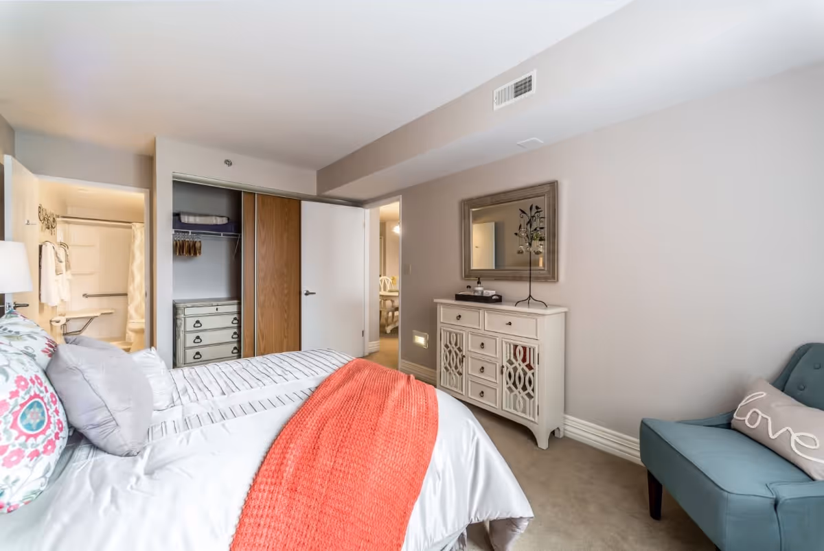 A cozy bedroom in Gambrill Gardens Senior Apartments featuring a neatly made bed with white and floral bedding and a coral throw blanket. There is a white dresser with a decorative mirror above it, a teal armchair with a pillow that says 'love', and an open closet with sliding doors. The bathroom is visible through an open door, showing a shower with grab bars and towels hanging on the wall.