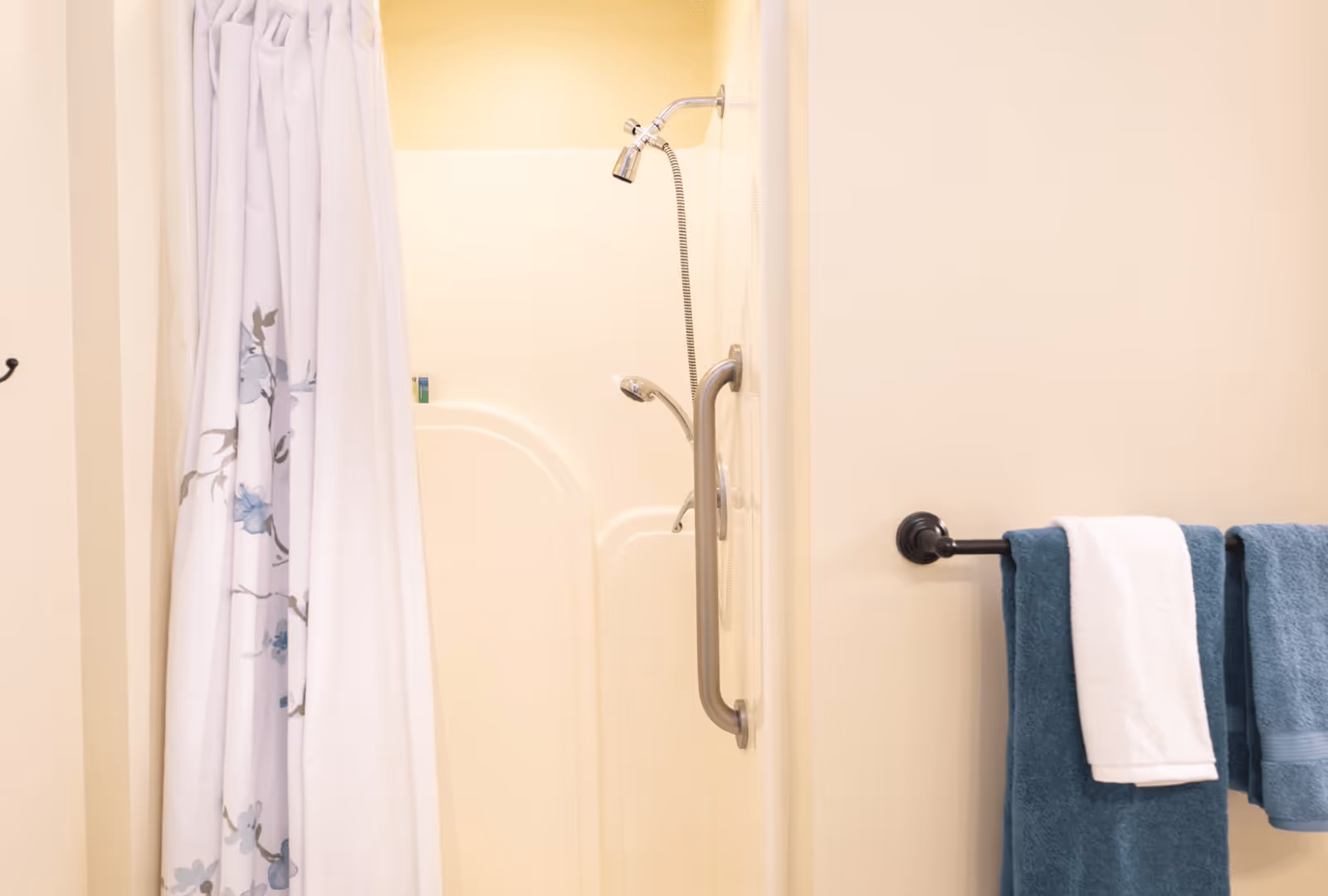 A bathroom shower area with a white and floral shower curtain partially drawn to the side. The shower has a handheld showerhead attached to a flexible hose and a grab bar for support. On the right wall, there is a towel rack holding two blue towels and one white towel.
