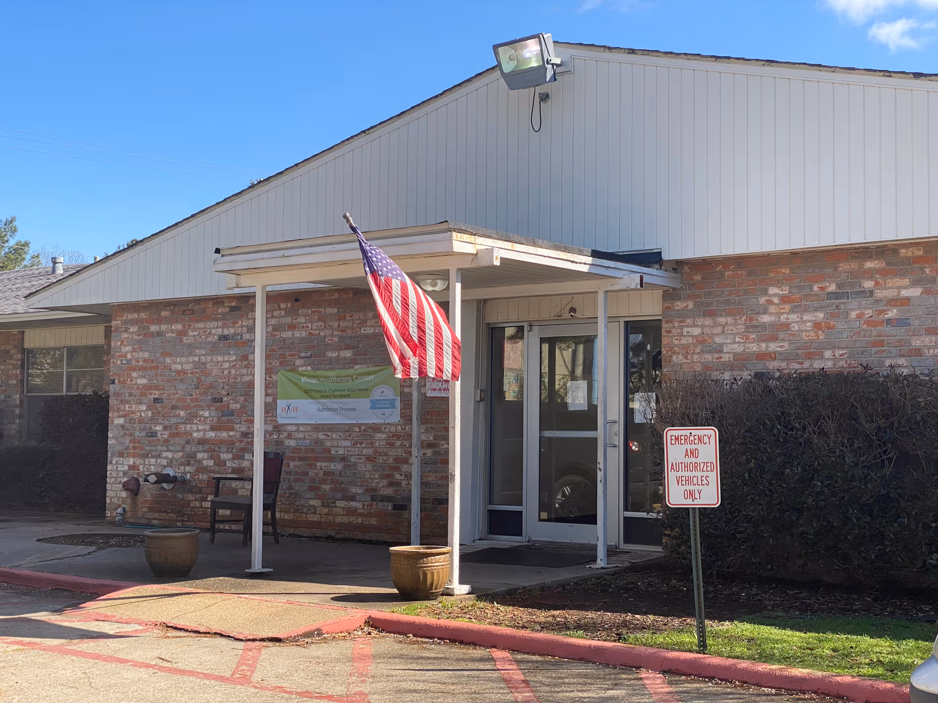 Exterior view of the entrance to Vivian Healthcare Center, a single-story brick building with a white overhang supported by two white pillars. An American flag is hanging from one of the pillars. There are two large planters near the entrance and a sign that reads 'EMERGENCY AND AUTHORIZED VEHICLES ONLY' near some bushes. The sky is clear and blue.