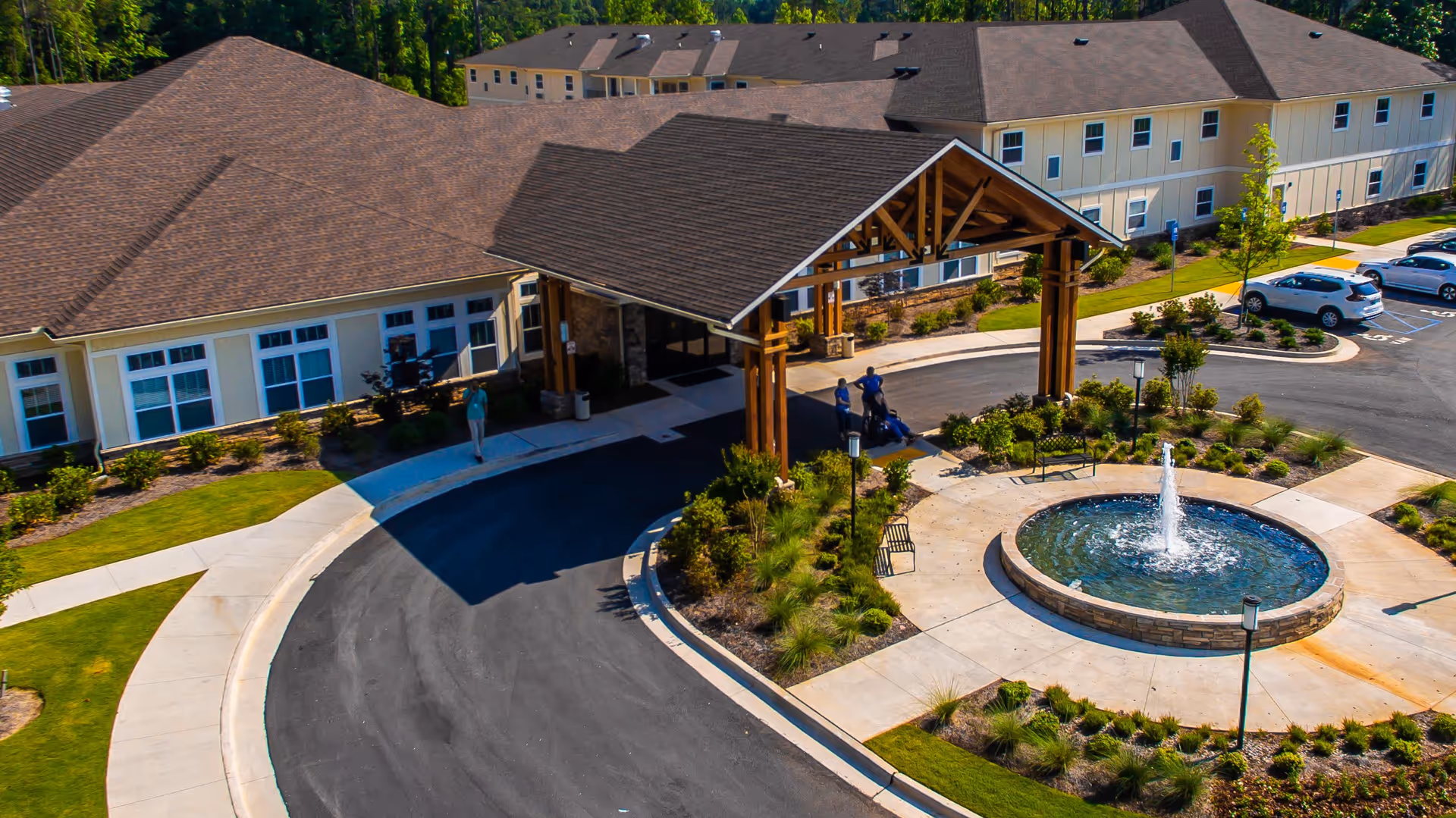 Aerial view of the entrance to Oaks at Oakland Plantation, showing a large covered drop-off area supported by wooden beams, a circular driveway, landscaped gardens, a round fountain with water spraying upwards, benches, and several parked cars. The building is a two-story structure with beige siding and multiple windows.