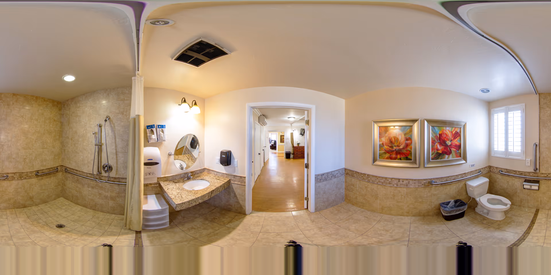 A spacious, accessible bathroom with beige tiled walls and floor. The bathroom features a walk-in shower with a curtain, a wall-mounted showerhead, and grab bars. There is a granite countertop with a sink, a round mirror, and wall-mounted soap and paper towel dispensers. On the opposite side, there is a toilet with grab bars, a trash bin, and two framed floral paintings on the wall. A window with white shutters allows natural light into the room. The bathroom opens into a hallway with wooden flooring and additional rooms visible in the distance.