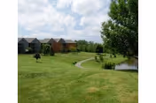 Wide green lawn with a winding path, a small pond and trees leading to apartment-style buildings under a partly cloudy sky.