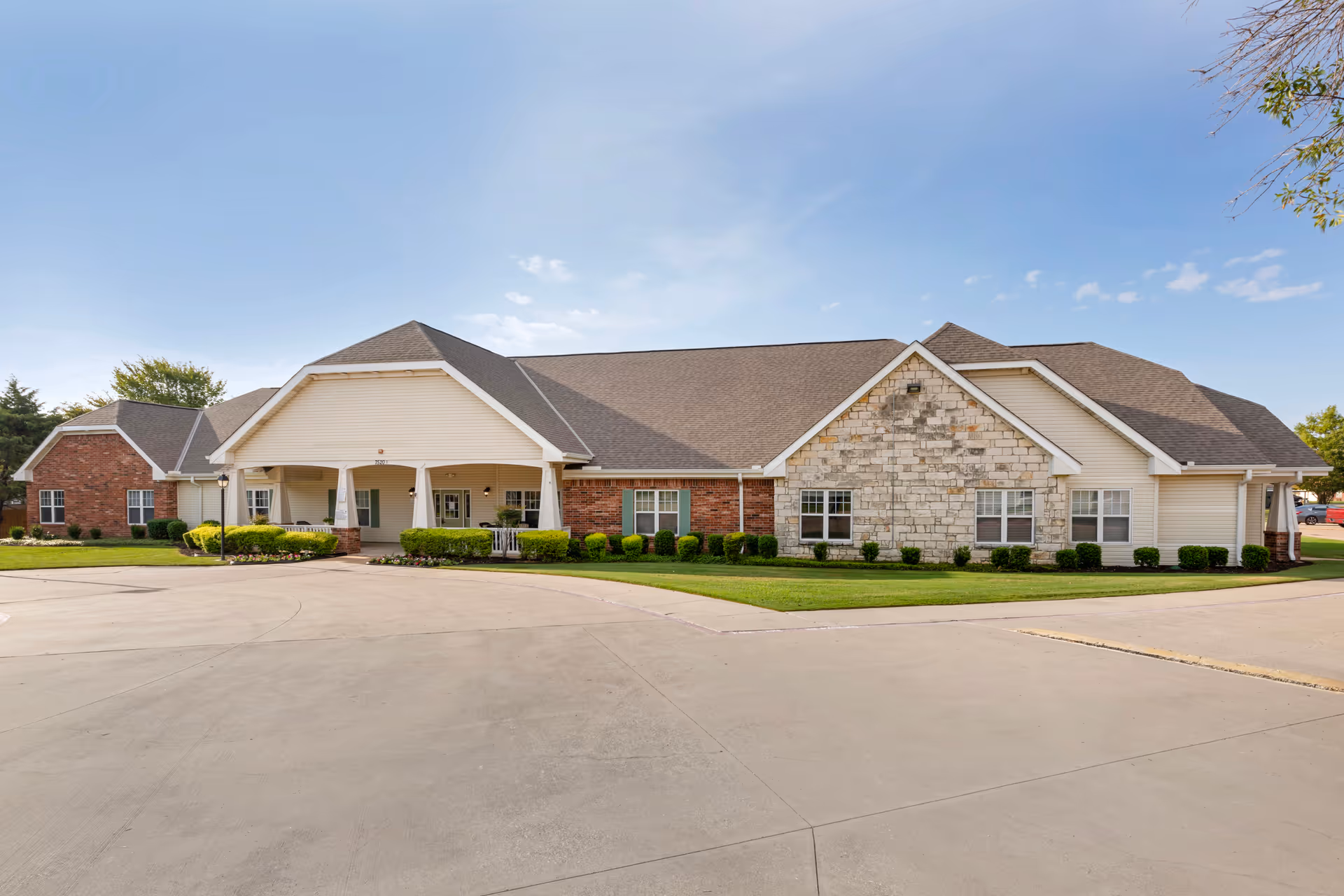 Exterior view of a single-story senior living facility building with a combination of brick, stone, and siding on the facade. The building has a covered entrance with white pillars and a driveway in front. There are neatly trimmed bushes and a well-maintained lawn surrounding the building under a clear blue sky.