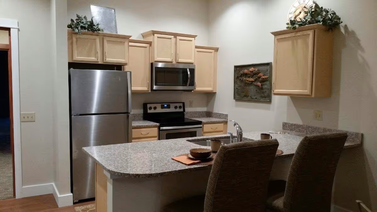 Kitchen with a granite countertop island, stainless steel refrigerator and stove, light wood cabinets, and two bar chairs.