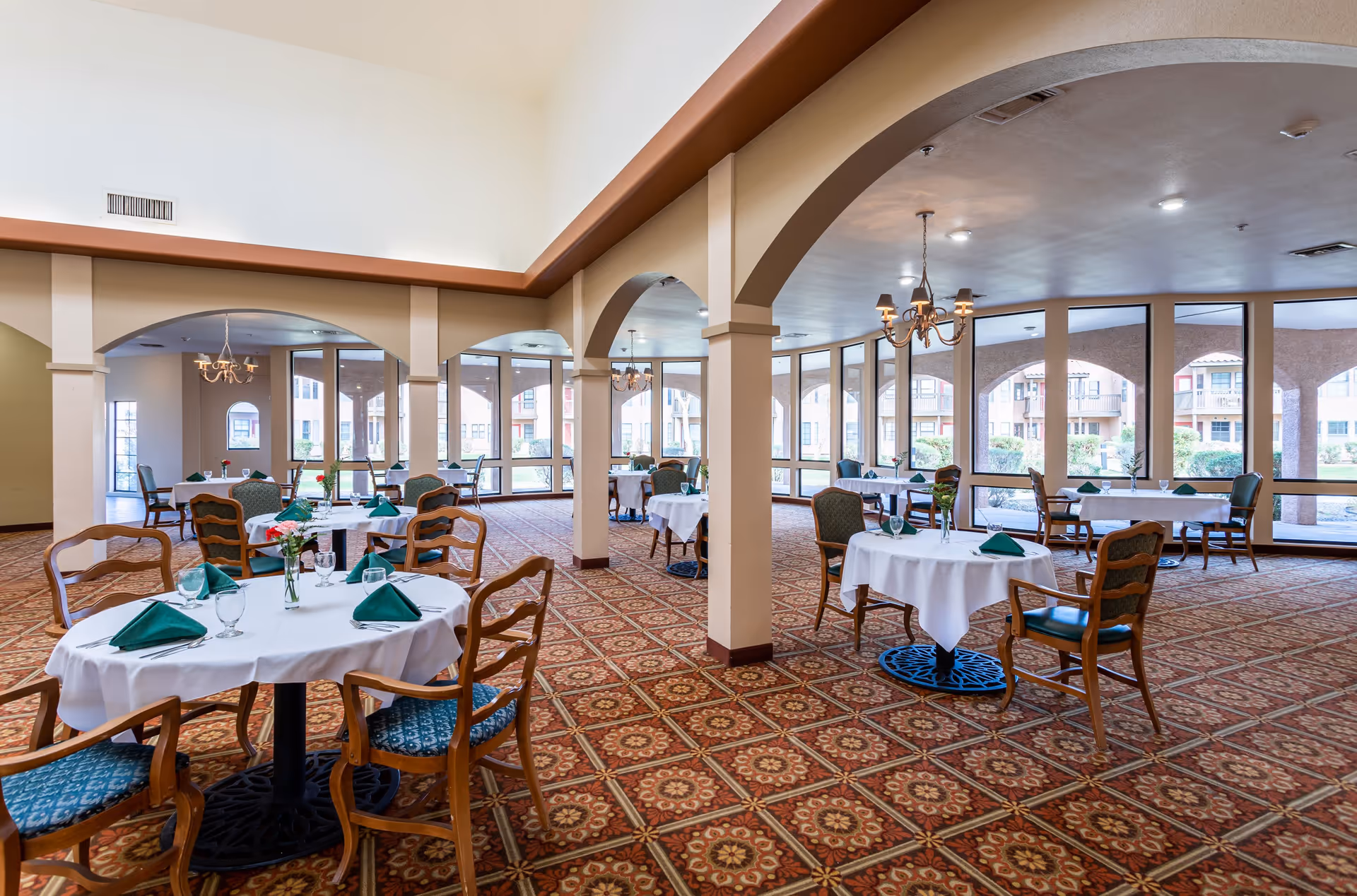 A spacious dining room with round tables covered in white tablecloths, each set with green folded napkins, glassware, and small floral centerpieces. The room features large windows with views of an outdoor courtyard, patterned carpet flooring, and arched architectural details.