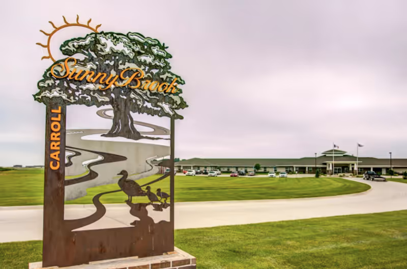Outdoor view of a senior living facility with a large green lawn and a curved driveway. In the foreground, there is a decorative metal sign featuring a tree, a winding path, and ducks, with the words 'SunnyBrook' and 'Carroll'. The building is visible in the background under a cloudy sky.