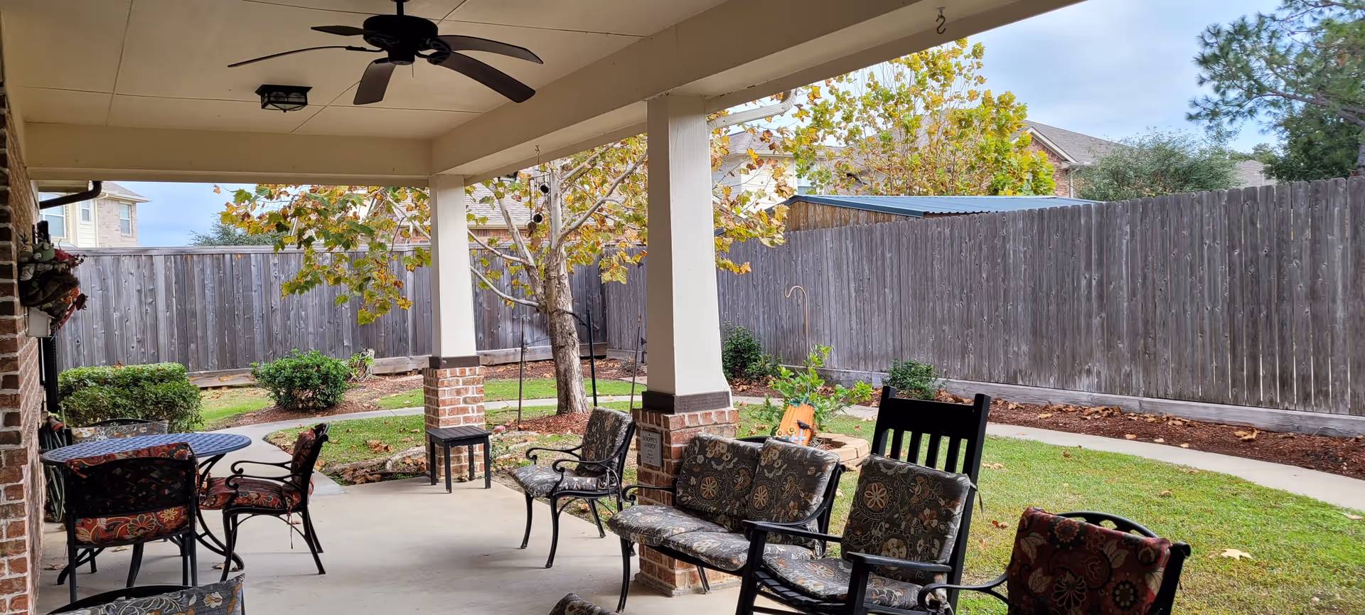 Covered outdoor patio area with ceiling fan, several cushioned chairs, a small table, and a view of a fenced backyard with a tree and shrubs.