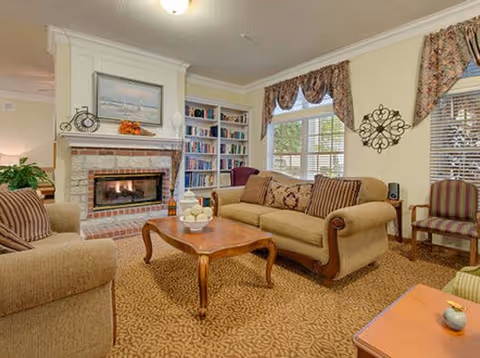 A cozy living room with a lit fireplace, beige sofas with patterned cushions, a wooden coffee table with decorative items, a bookshelf filled with books, and windows with floral curtains letting in natural light.