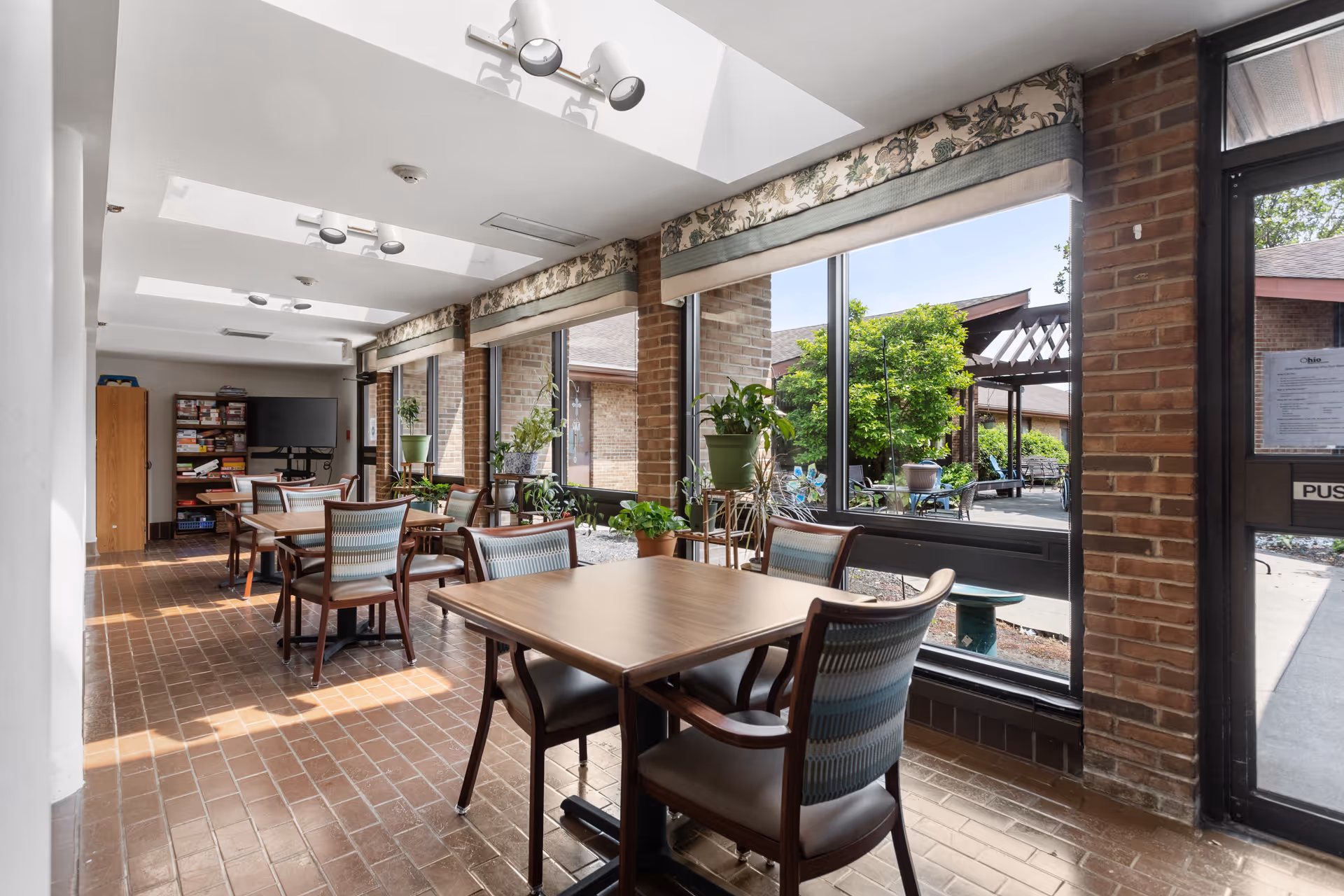 A bright common area with multiple tables and chairs arranged for seating. Large windows line one side, allowing natural light to fill the space and providing a view of an outdoor garden area with greenery and a pergola. Several potted plants are placed near the windows. The floor is tiled with brown bricks, and the ceiling has skylights and modern light fixtures. A TV and shelves with board games are visible in the background.