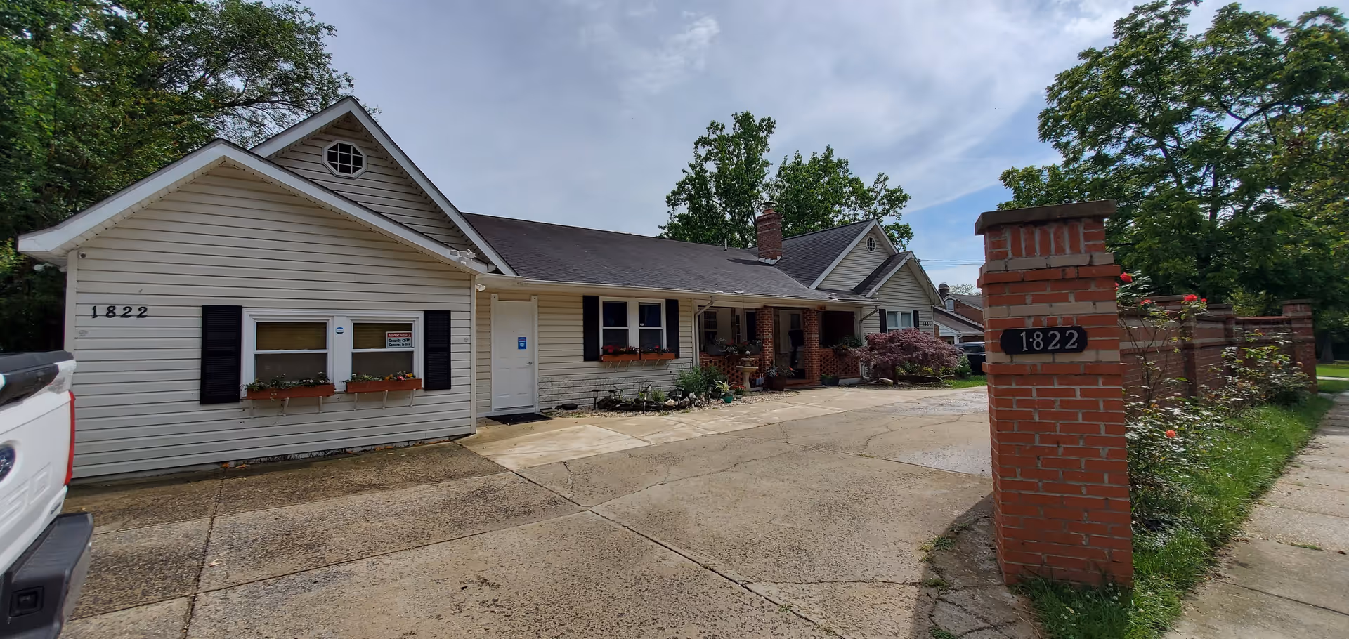 Exterior view of a single-story residential building with beige siding and black shutters, featuring flower boxes under the windows. The building has a driveway and a brick pillar with the number 1822. Trees and greenery surround the property under a partly cloudy sky.