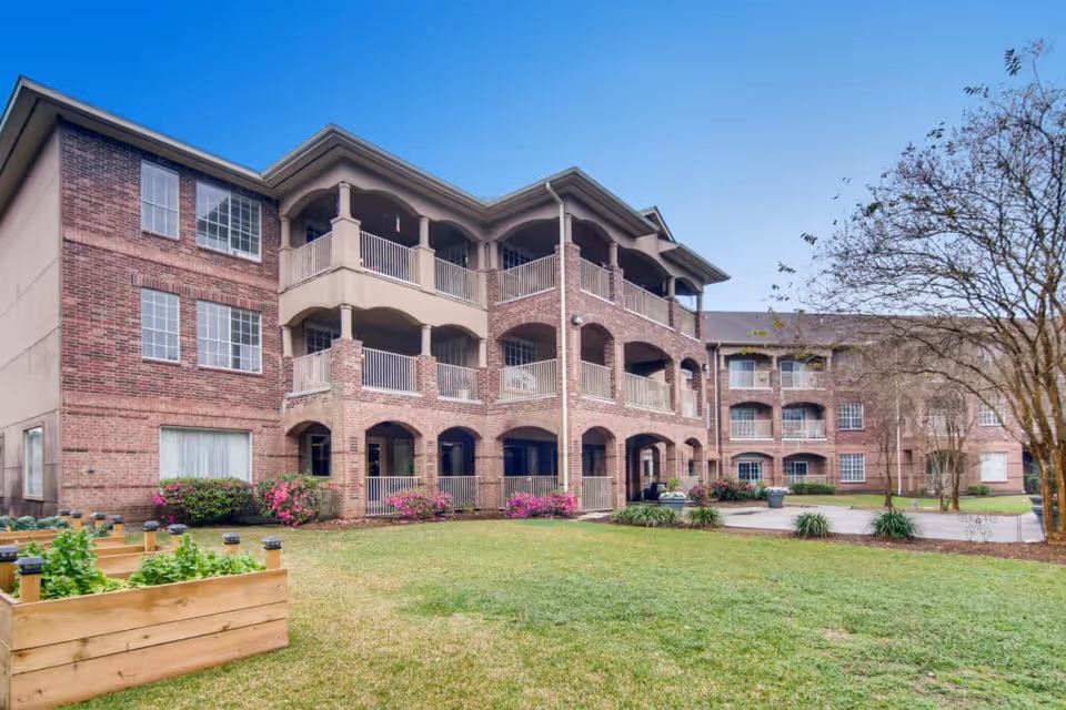Three-story brick senior living building with covered balconies overlooking a grassy courtyard and raised garden beds.
