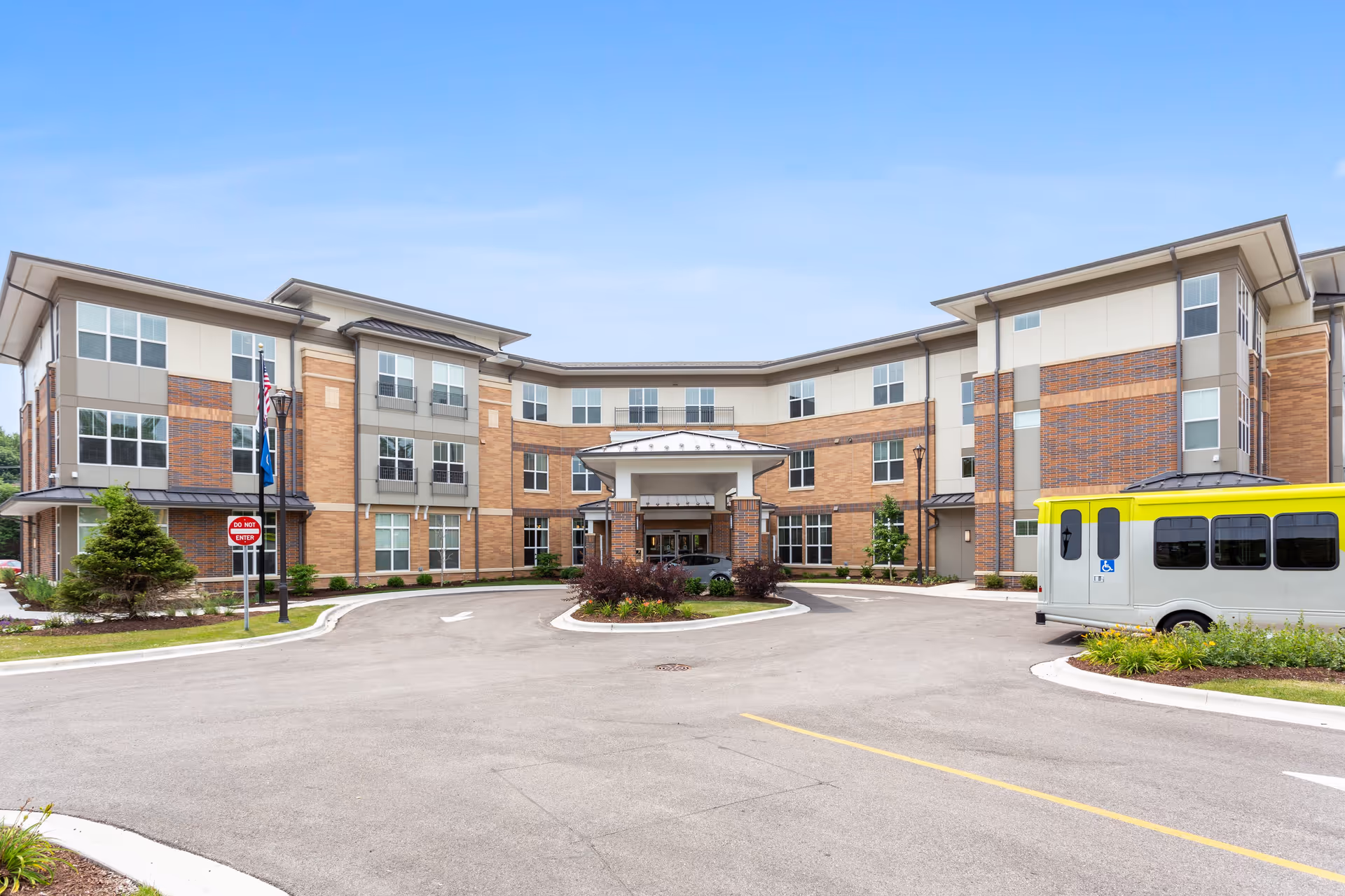 Front exterior view of a three-story senior living facility building with a covered entrance, multiple windows, and a driveway with a yellow and white shuttle bus parked on the right side. There are small landscaped areas with shrubs and trees around the building.
