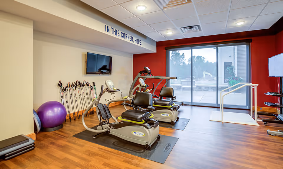 A fitness room with two NuStep exercise machines on mats, a purple exercise ball, several walking poles lined up against the wall, a wall-mounted TV, and a large window with a view of an outdoor patio. The wall above the TV has the phrase 'IN THIS CORNER, HOPE.'