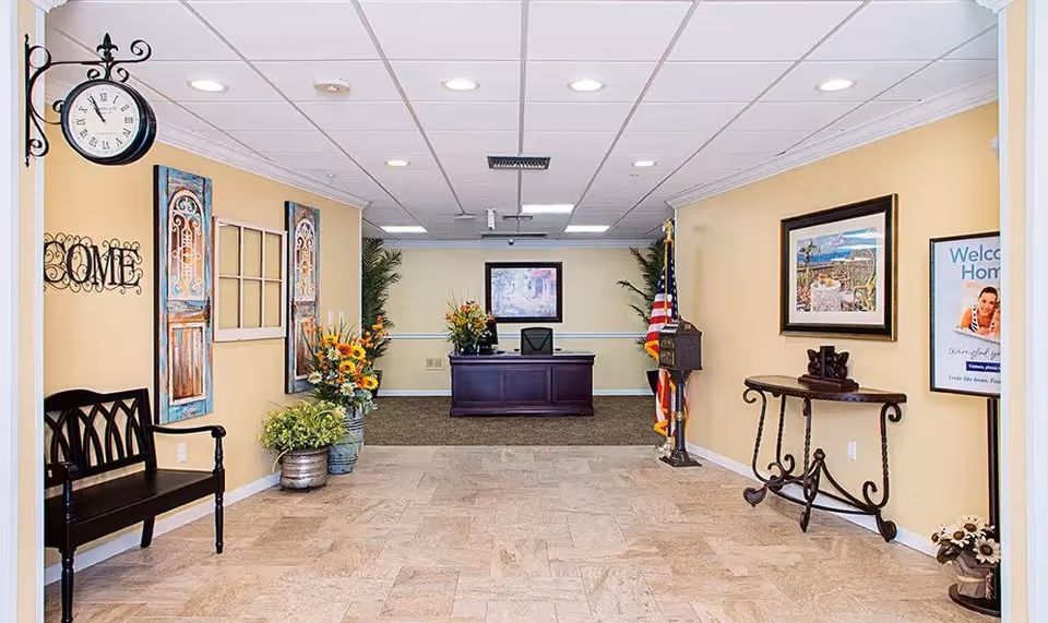 Reception area of a senior living facility with a dark wooden desk, an office chair, and decorative plants and flowers. The walls are painted light yellow and adorned with framed artwork and decorative items. There is a black bench on the left side and a small ornate table on the right side. An American flag stands near the desk, and a clock hangs on the left wall.