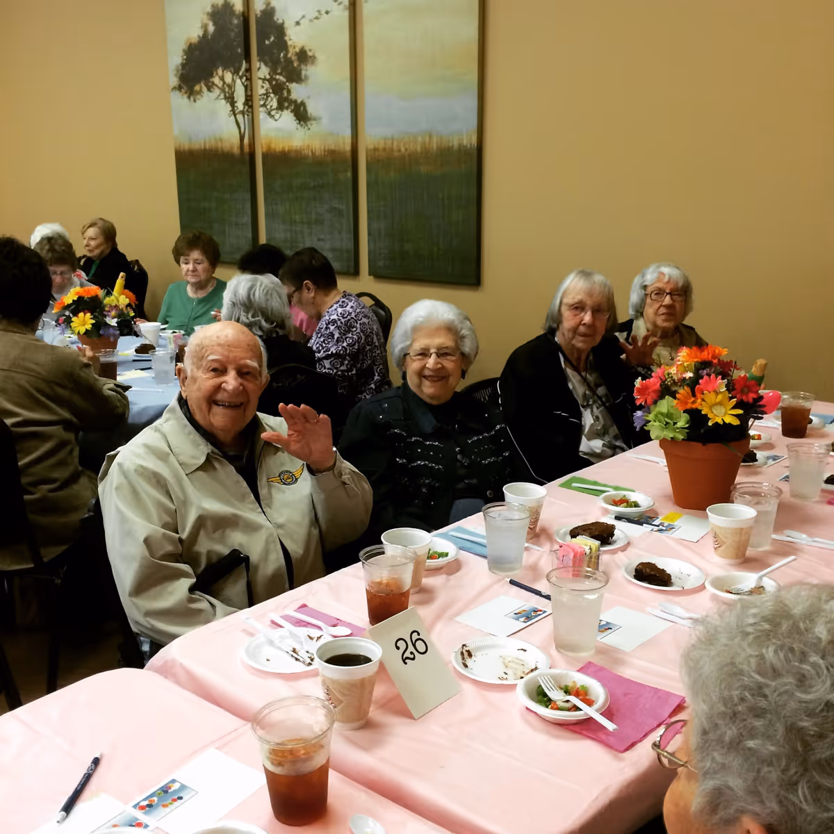 Elderly residents seated around a decorated long table in a dining room, smiling and eating.