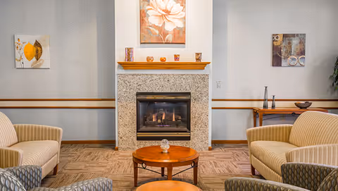 A cozy seating area with four upholstered armchairs arranged around two round wooden coffee tables in front of a lit fireplace with a granite surround. Above the fireplace is a floral painting, and on either side of the fireplace are decorative wall art pieces and a wooden console table with vases and a bowl.