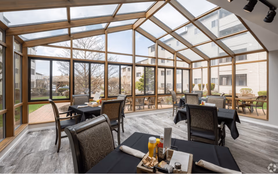 A bright dining area with large floor-to-ceiling windows and a glass ceiling, allowing natural light to fill the space. Several tables are set with black tablecloths, chairs with patterned upholstery, and condiments including ketchup and mustard. Outside, there is a view of a courtyard with trees and shrubs, and surrounding buildings.