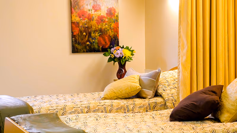 A cozy bedroom with two single beds covered in patterned bedspreads. Each bed has multiple pillows in shades of yellow, brown, and gray. A vase with colorful flowers sits on a nightstand between the beds. A painting of red and yellow flowers hangs on the wall above the nightstand. Yellow curtains are partially visible on the right side of the image.