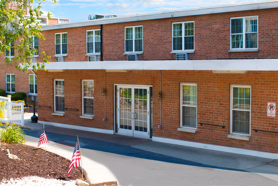 Brick exterior and main entrance of a senior living building with double glass doors, multiple windows, and small American flags in the landscaped foreground.