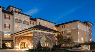 Exterior view of McDermott Crossing senior living facility at dusk, showing a multi-story building with warm lighting, a covered entrance with stone arches, and landscaped surroundings under a clear blue sky.
