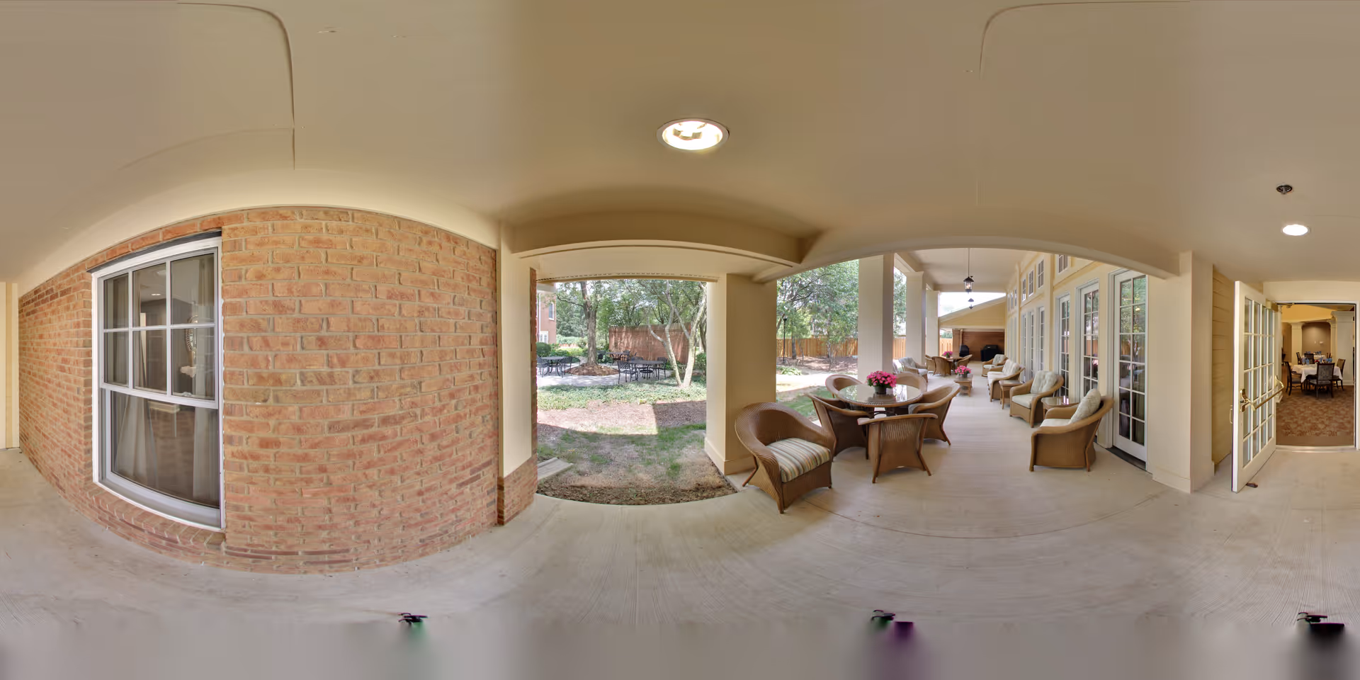 Covered outdoor patio area with wicker chairs and tables, some with flower arrangements, adjacent to a brick wall and large windows. The patio overlooks a garden with trees and a small seating area. There are ceiling lights and an open doorway leading to an interior room with dining tables and chairs.