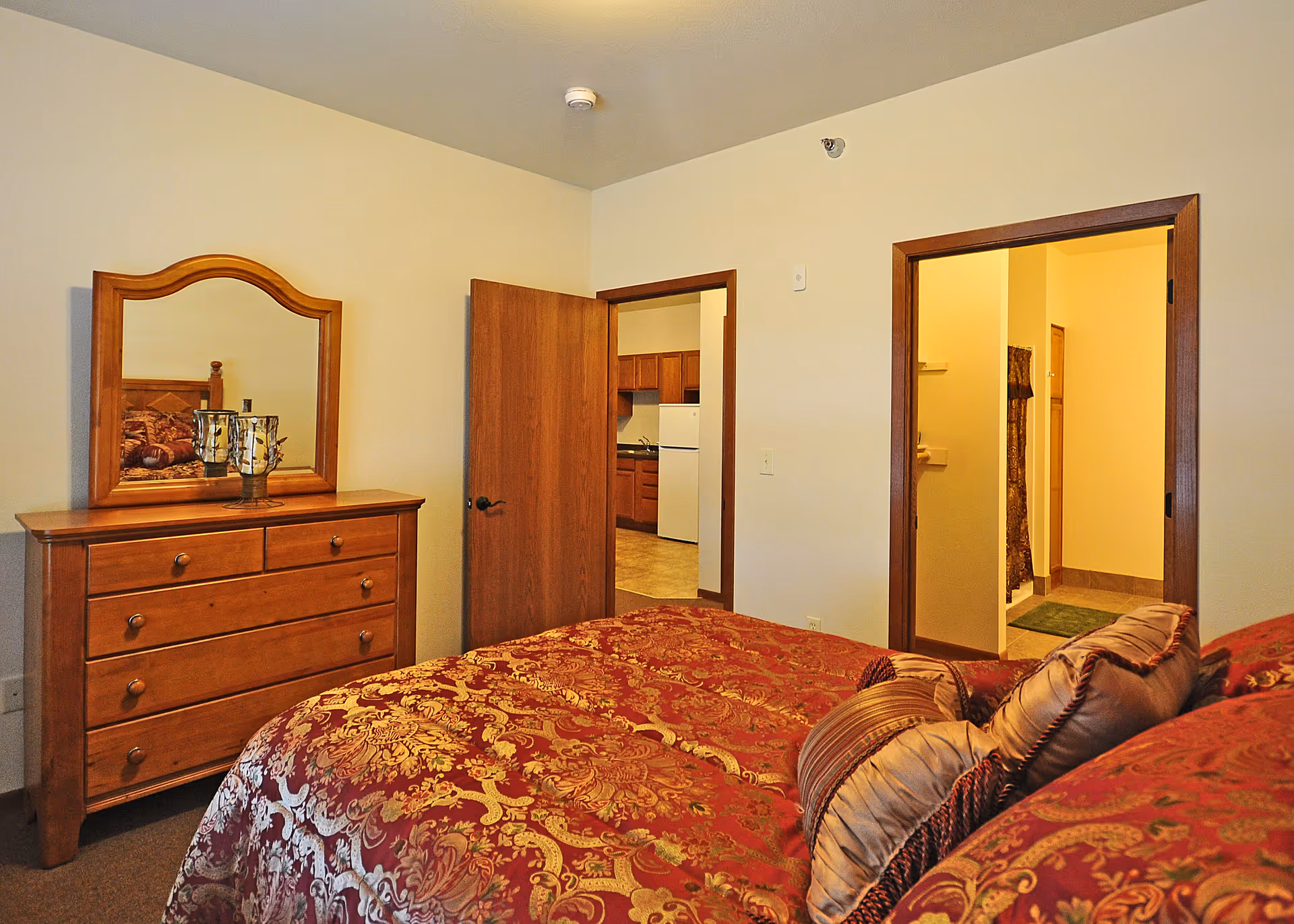 A bedroom with a wooden dresser and mirror on the left, a bed with a red and gold patterned comforter and multiple pillows in the foreground. Two doorways are visible, one leading to a kitchen area with wooden cabinets and a white refrigerator, and the other leading to a bathroom with a tiled floor and a shower curtain.