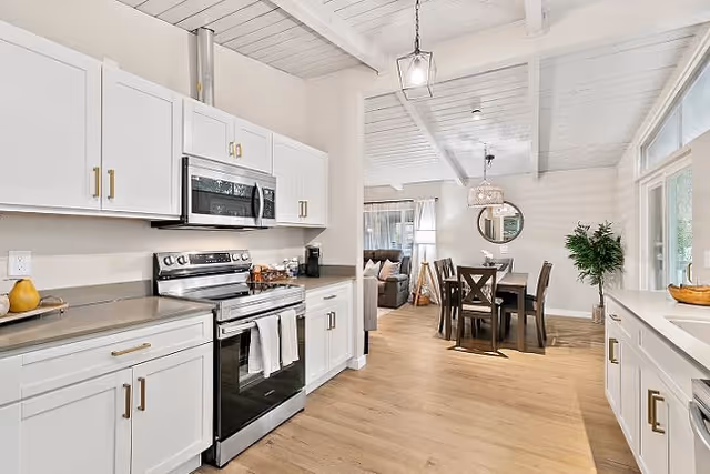 Bright and modern kitchen with white cabinets, stainless steel stove and microwave, light wood flooring, and a dining area with a wooden table and chairs in the background. The space has a clean, open layout with natural light coming through large windows.