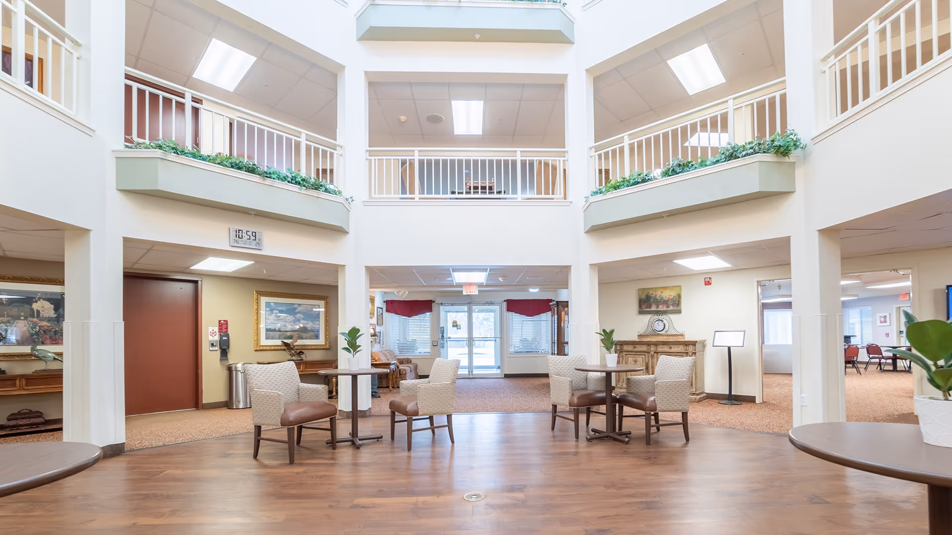 A spacious, well-lit common area inside a senior living facility with high ceilings and a second-floor balcony overlooking the space. The room features several small round tables with cushioned chairs, potted plants on the tables, and artwork on the walls. The flooring is a combination of wood and carpet, and there are large windows and glass doors letting in natural light.