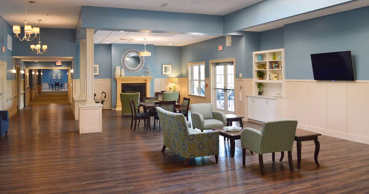 A spacious and well-lit common living area in Meadowview Memory Care facility featuring wooden flooring, blue walls with white wainscoting, a fireplace with decorative items and a round mirror above it, several armchairs and tables arranged for seating, a wall-mounted flat-screen TV, built-in shelves with books and decor, and a hallway leading to other rooms.