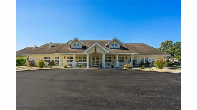 Front exterior view of a single-story building with a large paved driveway, a covered entrance with white columns, and an American flag hanging above the entrance. The building has a beige facade, multiple windows, and a sloped roof with dormer windows. There are small landscaped areas with shrubs and plants on either side of the entrance under a clear blue sky.
