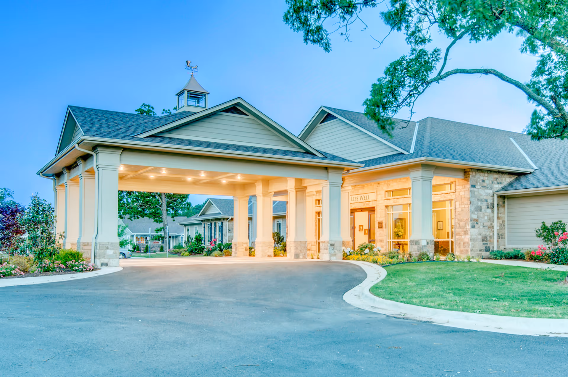 Exterior view of a senior living facility entrance with a covered driveway supported by columns, surrounded by landscaped greenery and trees under a clear blue sky.