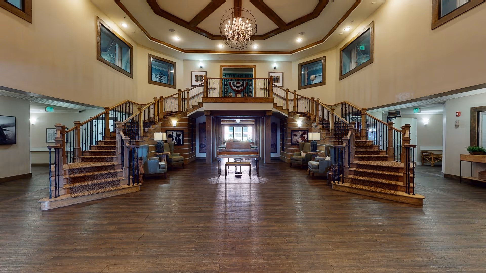 Spacious interior lobby area of a senior living facility with a grand double staircase featuring wooden railings and carpeted steps. The room has a high ceiling with a chandelier, several armchairs arranged along the walls, framed artwork, and large windows near the ceiling. The floor is wooden, and there is a piano with a bench in the center of the room.