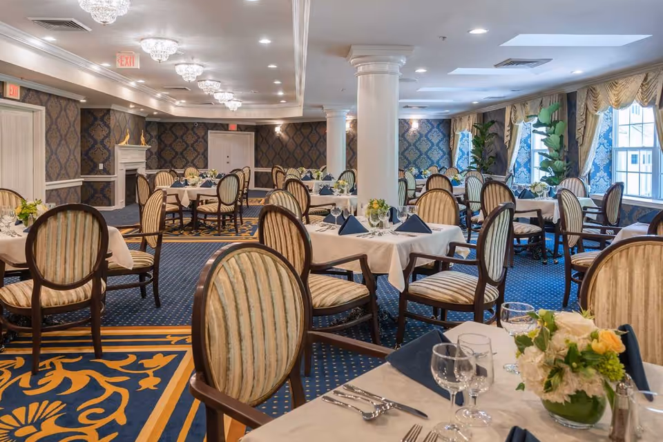 Elegant dining room with multiple tables covered in white tablecloths, set with glassware, silverware, and navy blue napkins. The room features patterned blue and gold carpet, striped upholstered chairs, decorative wallpaper, chandeliers, large windows with drapes, and potted plants near the windows.