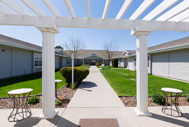 View through a white pergola structure looking down a concrete walkway flanked by green lawns and small bushes, leading to a single-story building with gray siding under a clear blue sky.