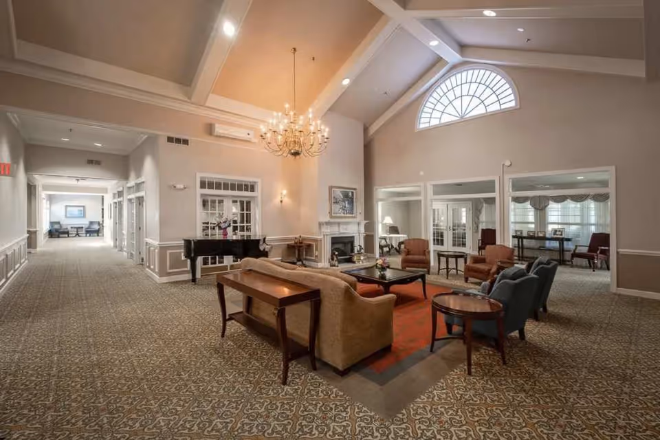 A spacious and elegant living room area in Chancellor's Village featuring a high vaulted ceiling with a large arched window, a chandelier, a fireplace, and comfortable seating including sofas and armchairs arranged around a coffee table on a patterned carpet. A black grand piano is positioned near the wall, and there are adjoining rooms visible through glass doors.