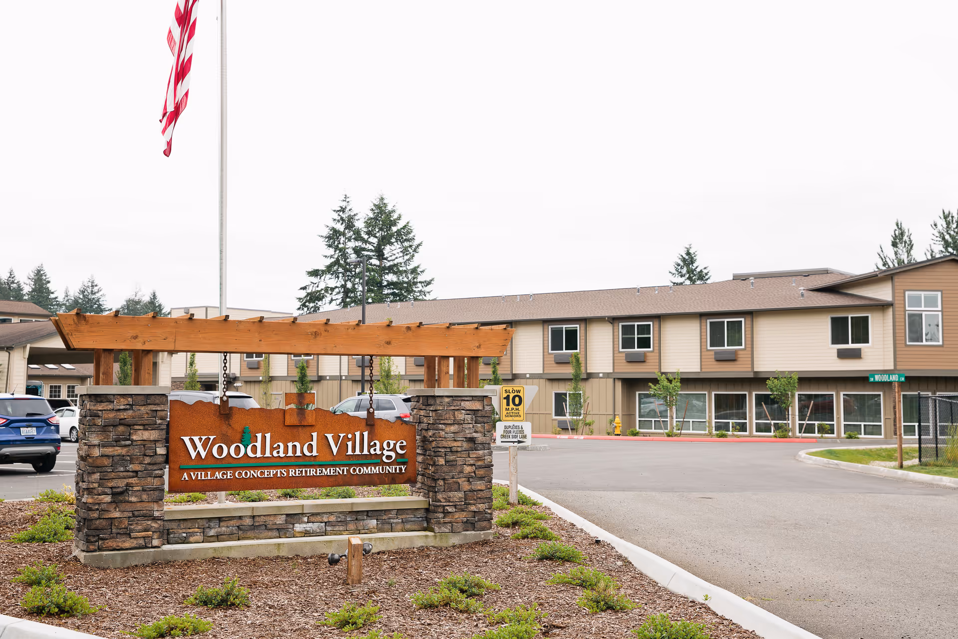 Woodland Village entrance sign with stone pillars in front of the retirement community building and a flagpole.