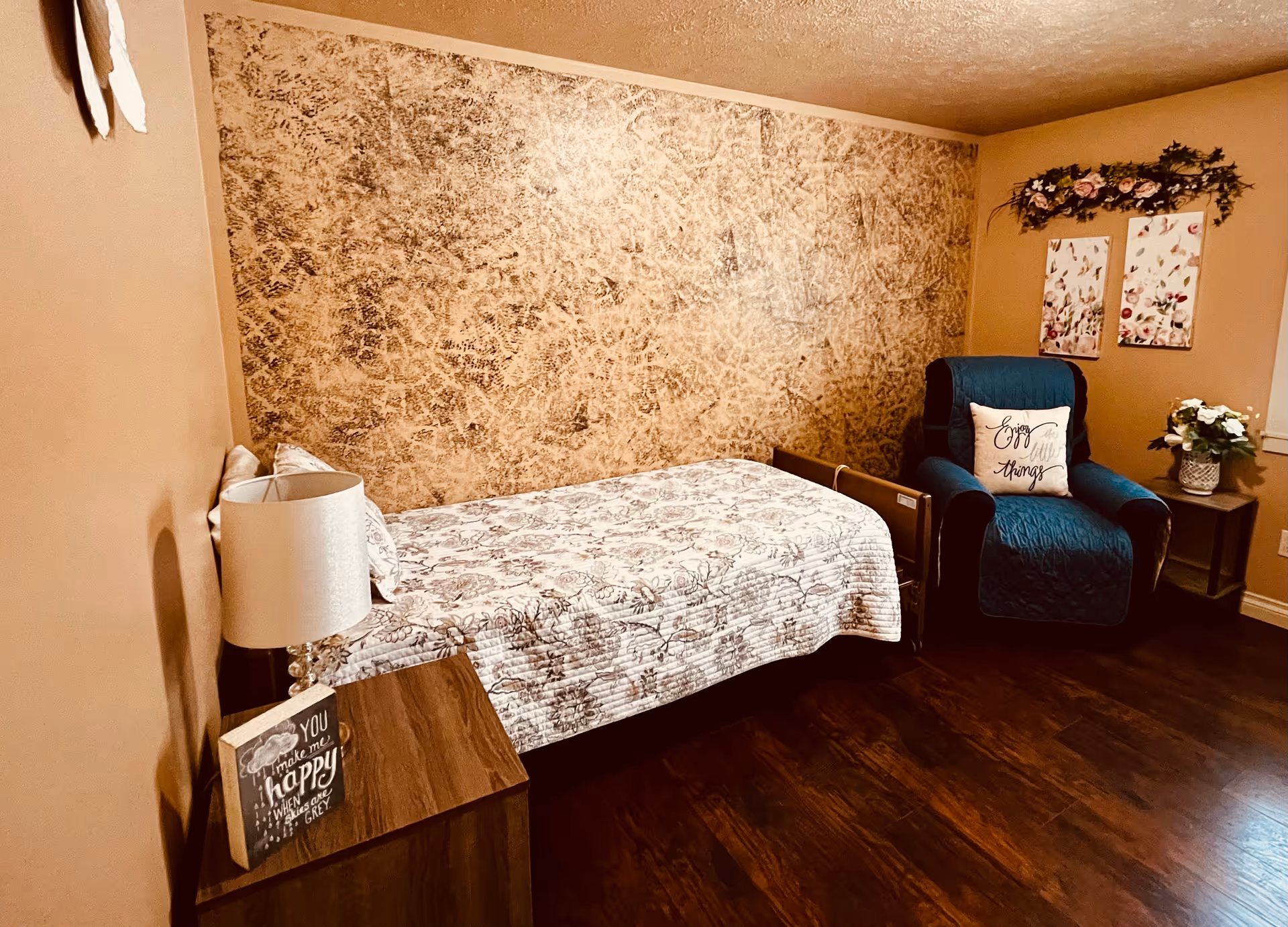 A cozy bedroom with a single bed covered in a floral quilt, a wooden nightstand with a lamp and a decorative sign, a blue armchair with a pillow that says 'Enjoy the little things', two floral wall art pieces, and a floral wreath above the chair. The room has warm beige walls and dark wooden flooring.