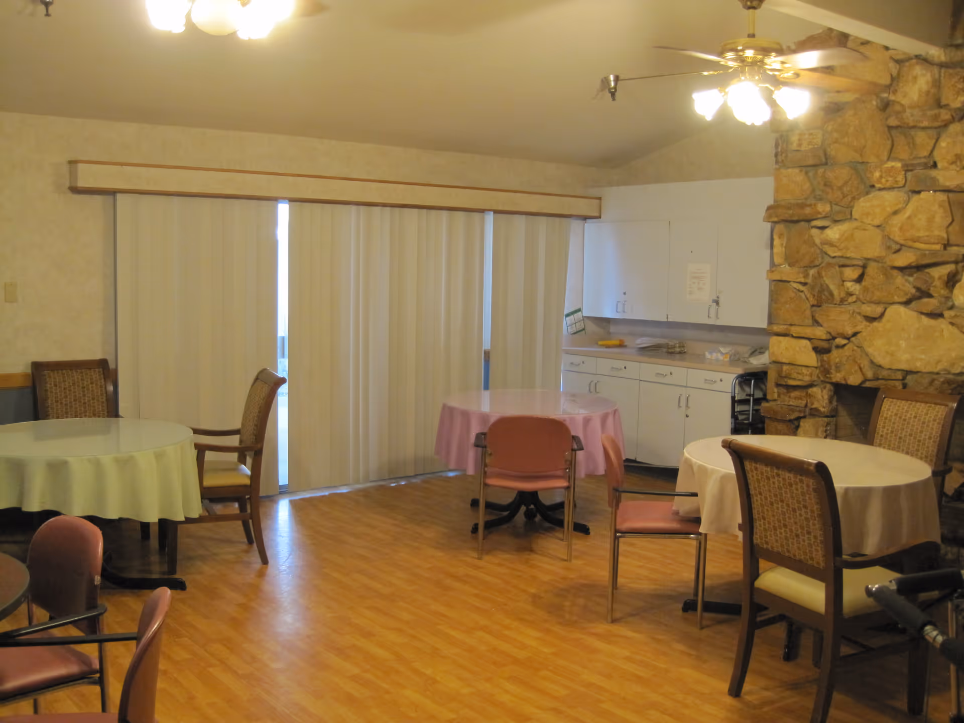 Interior view of a common dining area with round tables covered with tablecloths and surrounded by chairs. The room has wood flooring, a stone fireplace on the right, white cabinets along the back wall, and vertical blinds covering a sliding glass door.