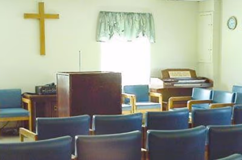 A small chapel or meeting room with rows of blue chairs facing a wooden podium. There is a wooden cross mounted on the wall to the left, a window with a green valance in the center, and an organ or piano against the right wall. The room has light-colored walls and a clock on the right side.