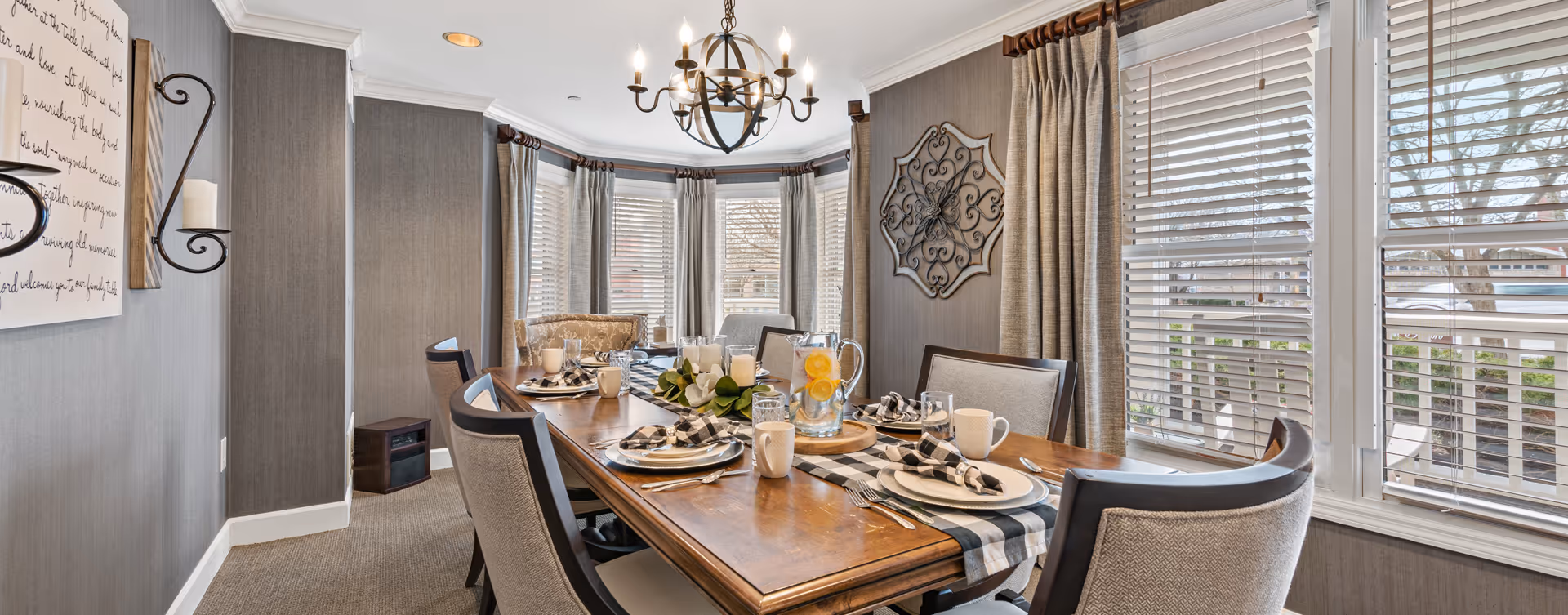 A well-lit dining room with a wooden dining table set for six people. The table is decorated with a black and white checkered runner, folded napkins, plates, glasses, mugs, and a pitcher of water with lemon slices. The room features large windows with white blinds and beige curtains, a decorative metal wall art piece, and a chandelier hanging from the ceiling. The walls are covered with textured gray wallpaper, and there is a cushioned bench in the corner.