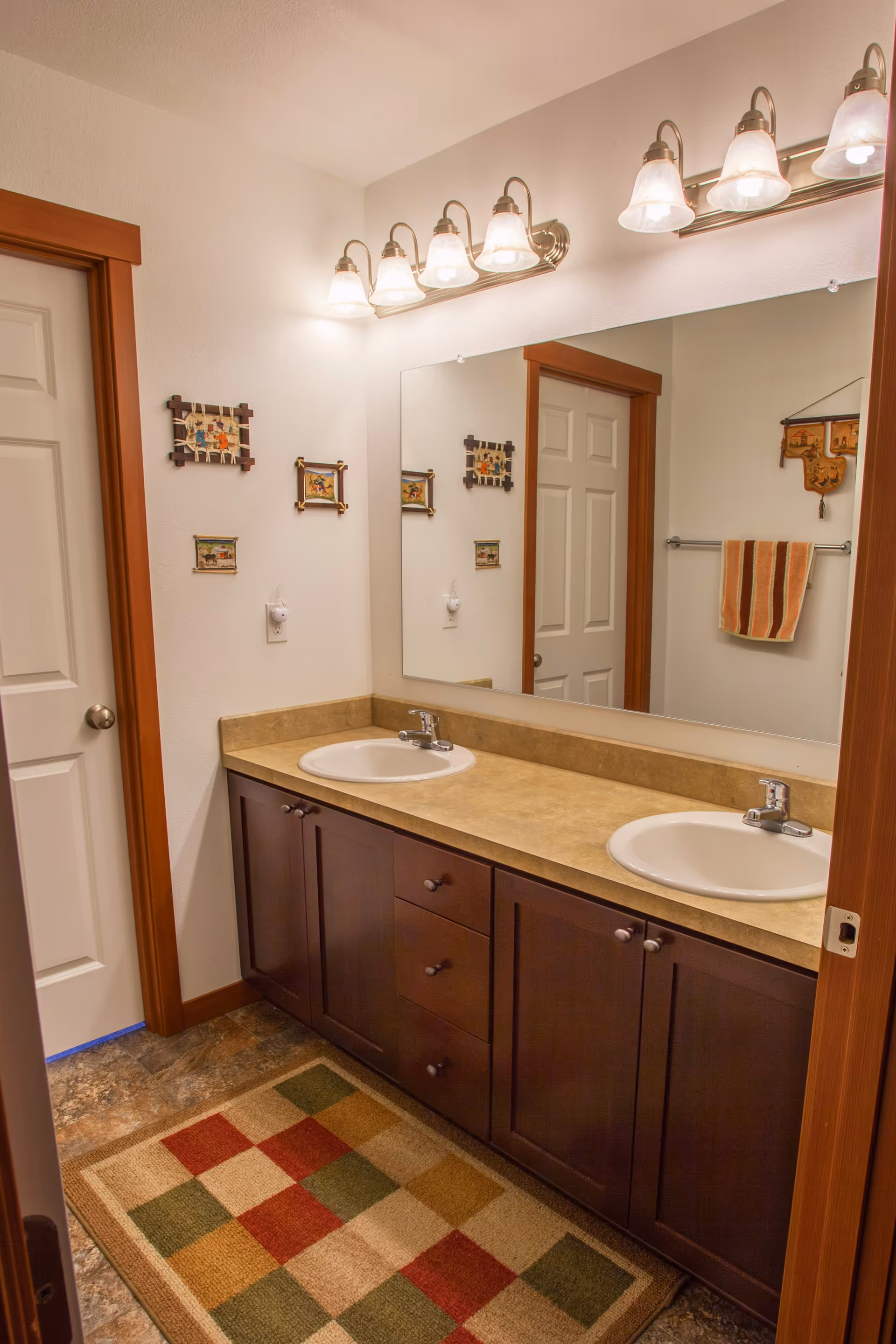 Bathroom with a double-sink vanity, large mirror, overhead light fixtures, decorative wall art and a patterned rug.